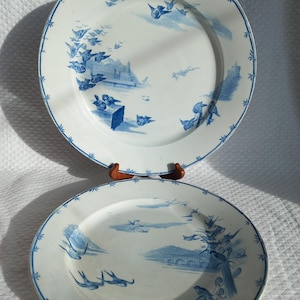 Peut inclure: Deux assiettes en céramique blanche avec des motifs bleus d'oiseaux et de paysages. Les assiettes ont un bord bleu avec un motif floral. Les assiettes sont présentées sur un support en bois.
