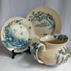 Puede incluir: Colección de vajilla de cerámica antigua de color crema con diseños florales y de pájaros azules. Incluye dos platos y una jarra. Los platos se exhiben sobre soportes negros. La jarra tiene un asa curva.