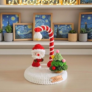 Pode incluir: Decoração de Natal de crochê com uma figura do Pai Natal, uma lâmpada em forma de bengala doce e uma pequena árvore de Natal. A base branca tem uma etiqueta de madeira que diz "Rose". O fundo inclui pinturas emolduradas e pequenas plantas em vasos.