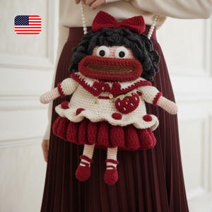 Könnte beinhalten: Handgefertigte Häkeltaschenpuppe in Form eines Mädchens, mit roter Schleife, schwarzen Haaren und Erdbeerapplikation. Die Tasche hat ein weißes und rotes Kleid und einen Perlenriemen.