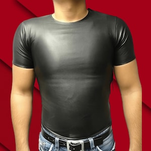 May include: Black leather-look t-shirt with short sleeves. The shirt has a crew neck and a form-fitting design. The model is wearing blue jeans and a black belt. The background is a gradient of red.