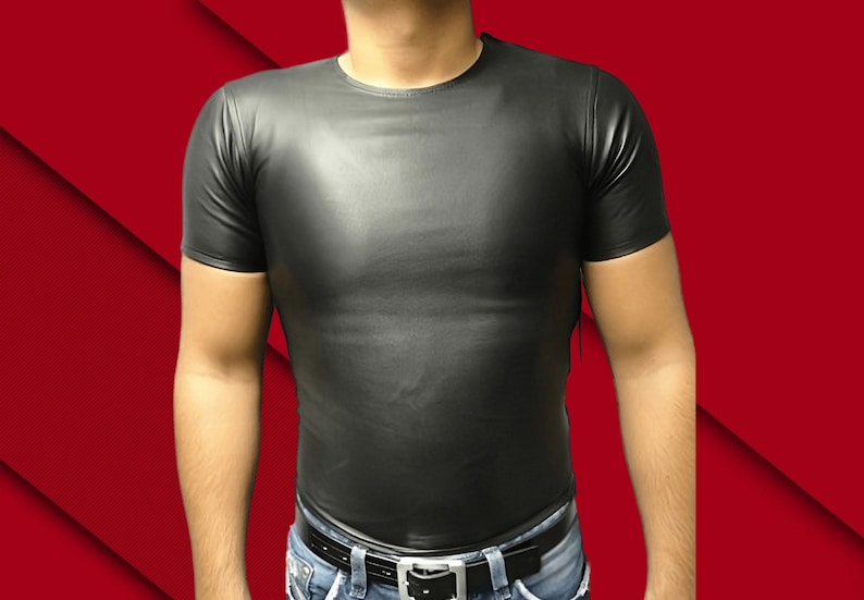 May include: Black leather-look t-shirt with short sleeves. The shirt has a crew neck and a form-fitting design. The model is wearing blue jeans and a black belt. The background is a gradient of red.