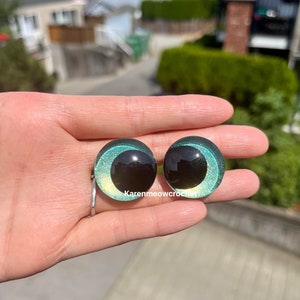 K&ouml;nnte beinhalten: Zwei runde, gr&uuml;ne und schwarze Kunststoffaugen mit Glitzer. Die Augen haben eine schwarze Pupille und eine gr&uuml;ne Iris mit goldenem Glitzer.