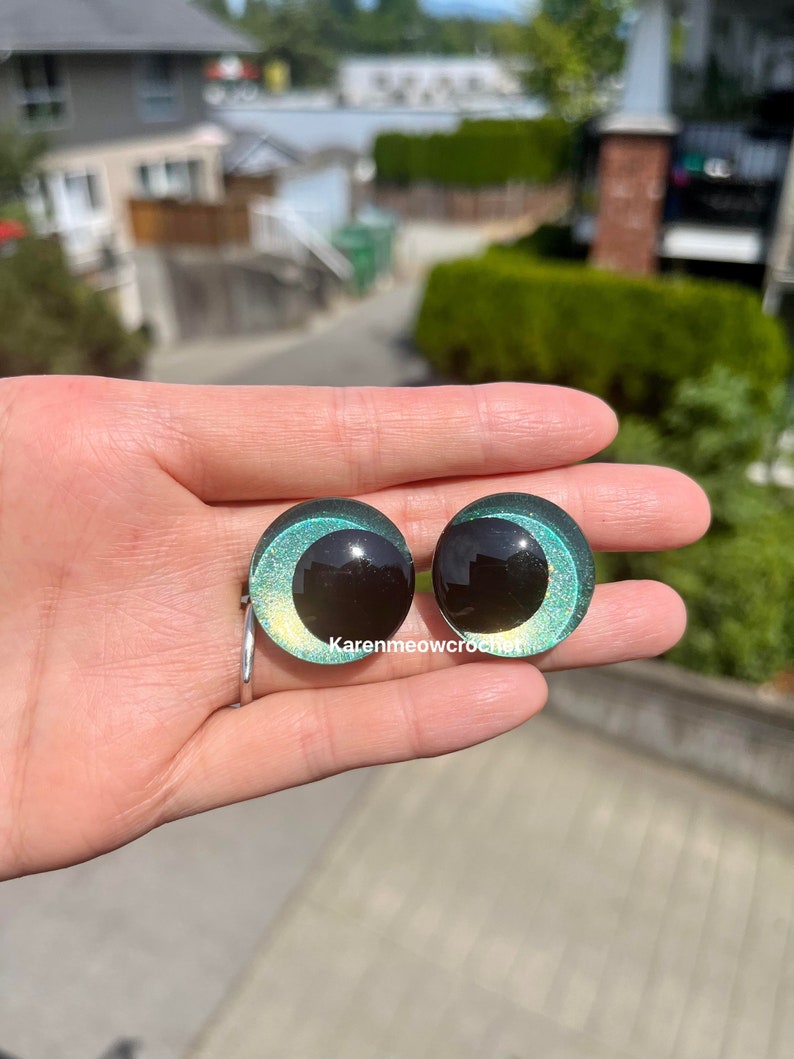 K&ouml;nnte beinhalten: Zwei runde, gr&uuml;ne und schwarze Kunststoffaugen mit Glitzer. Die Augen haben eine schwarze Pupille und eine gr&uuml;ne Iris mit goldenem Glitzer.