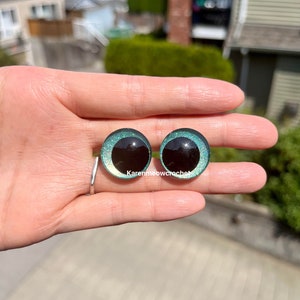 K&ouml;nnte beinhalten: Zwei runde, gr&uuml;ne, glitzernde Sicherheitsaugen mit schwarzen Pupillen. Die Augen werden in einer Hand gehalten.