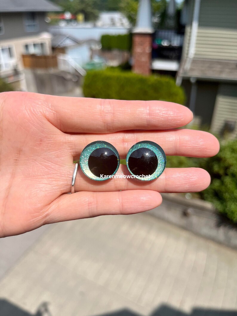 K&ouml;nnte beinhalten: Zwei runde, gr&uuml;ne, glitzernde Sicherheitsaugen mit schwarzen Pupillen. Die Augen werden in einer Hand gehalten.