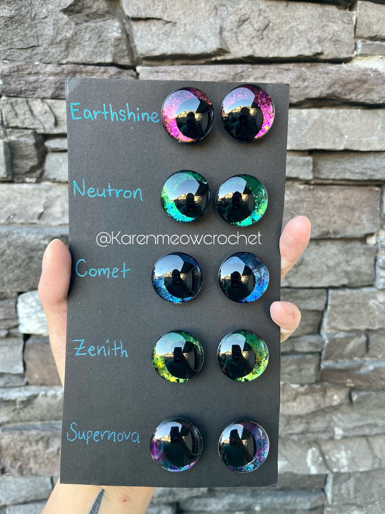 May include: A black card with five rows of colourful glass eyes. Each row is labelled with a name: Earthsine, Neutron, Comet, Zenith, and Supernova. The eyes are all different colours and have a glittery finish.