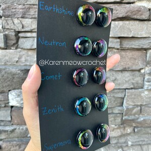 May include: A black card with the words "Earthshine", "Neutron", "Comet", "Zenith", and "Supernova" written in blue ink. Each word is paired with two round, iridescent, black resin buttons with a glittery, rainbow-like effect.