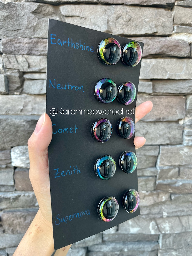 May include: A black card with the words "Earthshine", "Neutron", "Comet", "Zenith", and "Supernova" written in blue ink. Each word is paired with two round, iridescent, black resin buttons with a glittery, rainbow-like effect.