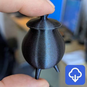 May include: A small, black cauldron with a lid and three legs. The cauldron is made of a smooth, shiny material.