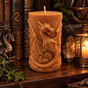 May include: A cylindrical pillar candle in a warm tan colour featuring a detailed, raised relief sculpture of a dragon with spread wings and a coiled tail wrapped around the base. The candle has a single white wick centred on the flat top surface.