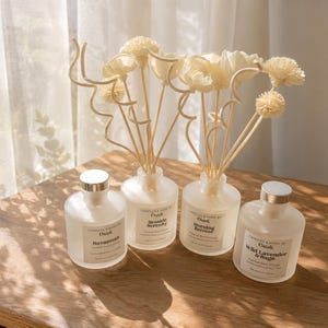 May include: Four frosted glass reed diffuser bottles with silver caps, each featuring a label reading Candles & More by Osadi. Two bottles contain decorative wooden sticks with flower-shaped tops, while all four display unique scent names.