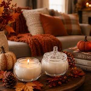 May include: Two glass pumpkin-shaped candles, one lit and one lidded, sit on a round wooden tray with a pinecone, faux autumn leaves, and a small orange pumpkin. Nearby are stacked books labelled AUTUMN, HOME, and living simply.