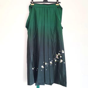Può includere: Una gonna hakama plissettata verde e nera con ricami floreali bianchi. La gonna è appesa a una gruccia nera su sfondo bianco. La gonna ha un effetto sfumato, che passa dal verde al nero.