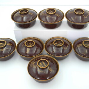 May include: Set of seven brown and gold ceramic bowls with a traditional Japanese design. The bowls have a raised rim and a flat bottom. The design features a stylized landscape with mountains, trees, and water.