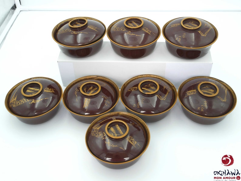 May include: Set of seven brown and gold ceramic bowls with a traditional Japanese design. The bowls have a raised rim and a flat bottom. The design features a stylized landscape with mountains, trees, and water.