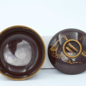 May include: A set of two round, dark brown lacquer bowls with gold accents. The lid features a stylized mountain scene with clouds and birds.