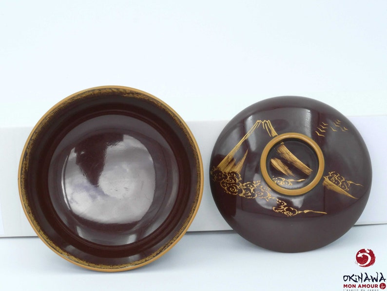 May include: A set of two round, dark brown lacquer bowls with gold accents. The lid features a stylized mountain scene with clouds and birds.