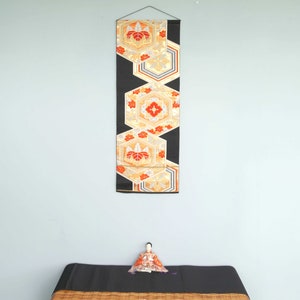 May include: A black and gold Japanese wall hanging with a geometric pattern of hexagons and red flowers. The wall hanging is hanging on a light blue wall.