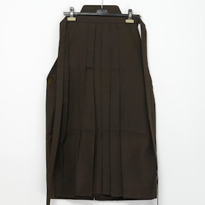 Könnte beinhalten: Ein dunkelbrauner Hakama, ein traditionelles japanisches Kleidungsstück, hängt an einem schwarzen Kleiderbügel vor weißem Hintergrund. Der Hakama hat tiefe Falten und ein strukturiertes Design. Das Kleidungsstück ist aus robustem Stoff gefertigt.