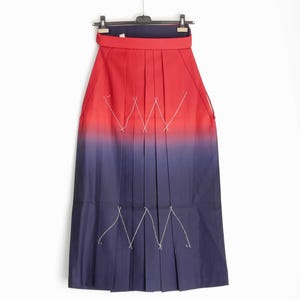 May include: A pleated skirt with a red to blue gradient. White zigzag stitching accents the front. The skirt is hanging on a black hanger against a white background. The skirt has a wide waistband.