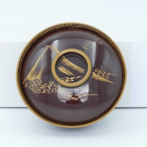 May include: A dark brown lacquer bowl with a gold rim and a design of a mountain and clouds. The bowl is decorated with gold paint.
