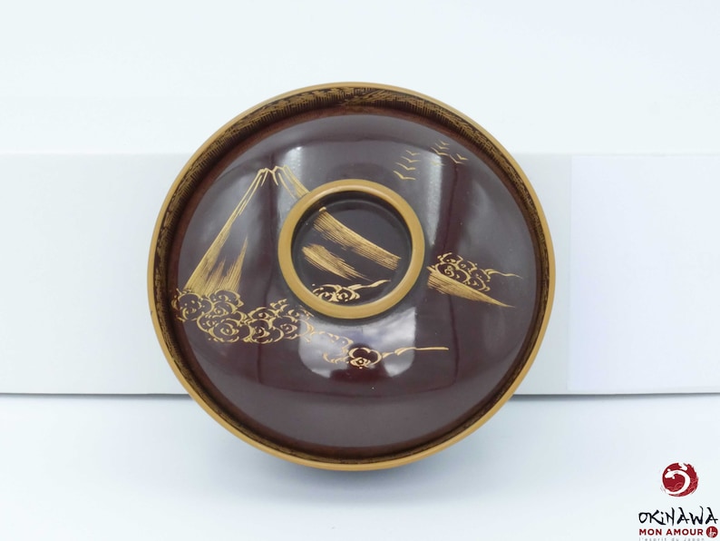 May include: A dark brown lacquer bowl with a gold rim and a design of a mountain and clouds. The bowl is decorated with gold paint.