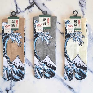 Op de afbeelding: Drie paar tabi-sokken met een Japans golfontwerp. De sokken zijn beige, grijs en wit. De sokken hebben de tekst "足袋" op het etiket.