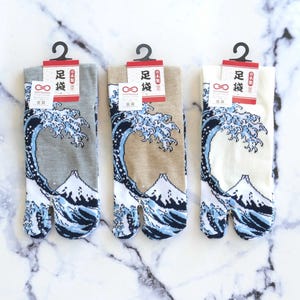 May include: Three pairs of tabi socks featuring a Japanese wave design. The socks are grey, beige, and white. Each sock has the text "足袋" in red on the tag.