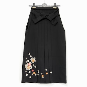 May include: Black hakama skirt with pleats, hanging on a hanger. The skirt features a floral design in white, orange, and pink near the bottom. The skirt is a traditional Japanese garment.