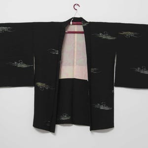 K&ouml;nnte beinhalten: Ein schwarzer japanischer Hanten, oder kurzer Kimono, mit einem wei&szlig;en und gr&uuml;nen Blumenmuster. Der Hanten h&auml;ngt an einem Holzb&uuml;gel.