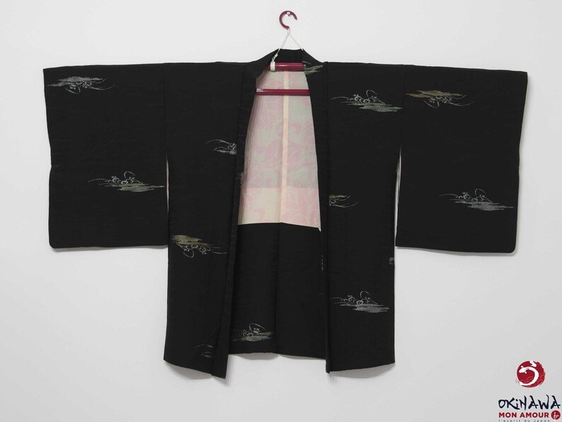 K&ouml;nnte beinhalten: Ein schwarzer japanischer Hanten, oder kurzer Kimono, mit einem wei&szlig;en und gr&uuml;nen Blumenmuster. Der Hanten h&auml;ngt an einem Holzb&uuml;gel.