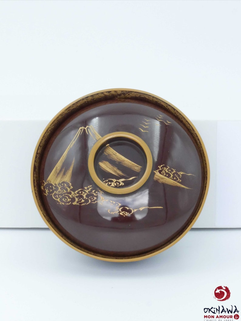 May include: A dark red Japanese bowl with a gold rim and a gold design of a mountain and clouds. The bowl is made of lacquered wood.