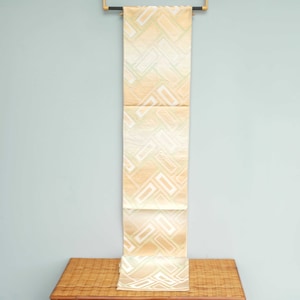 May include: A long, cream-colored fabric with a geometric pattern of squares and lines in a lighter shade of green. The fabric is hanging from a wooden rod against a light blue wall.