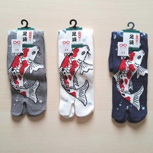 Può includere: Tre paia di calzini tabi, ognuno con un disegno di carpa koi in rosso, bianco e nero. I calzini sono grigi, bianchi e blu navy. Ogni paio ha un'etichetta con testo giapponese e un gancio nero.