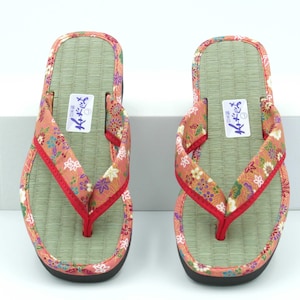 May include: A pair of red and green floral print Japanese sandals with a straw sole. The sandals have a red strap and a black sole.