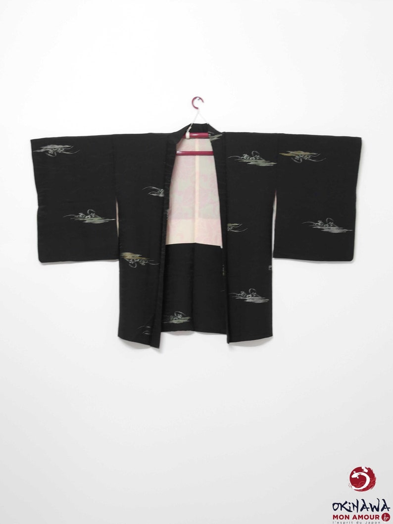 K&ouml;nnte beinhalten: Ein schwarzer Kimono mit einem wei&szlig;en und goldenen Muster aus stilisierten V&ouml;geln und Wellen. Der Kimono h&auml;ngt an einem Holzb&uuml;gel vor einem wei&szlig;en Hintergrund.