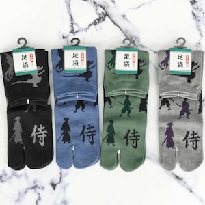 Può includere: Quattro paia di calzini tabi neri, blu, verdi e grigi con una grafica di un guerriero samurai e il testo "侍" sul fondo dei calzini.