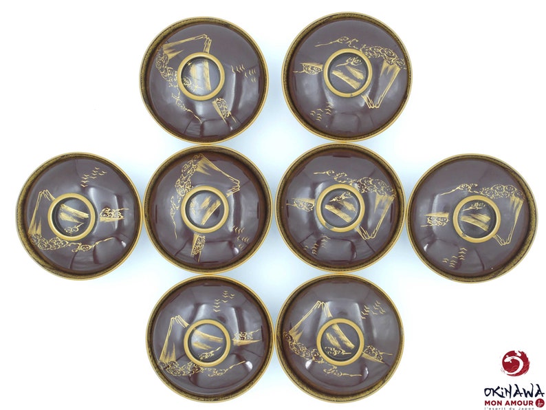 May include: Set of eight small, round, brown bowls with gold painted designs. Each bowl features a mountain scene with a bridge and trees. The bowls are arranged in a circle.