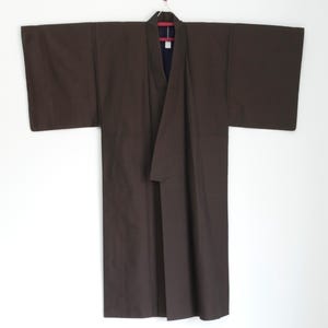 Könnte beinhalten: Ein brauner Kimono hängt vor einem weißen Hintergrund. Das traditionelle japanische Kleidungsstück hat weite Ärmel und eine Wickel-Front. Der Kimono ist aus einem einfarbigen, dunkelbraunen Stoff gefertigt und wird auf einem Kleiderbügel präsentiert.
