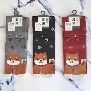 Può includere: Tre paia di calze a dita separate con un design di cane Shiba Inu. Le calze sono grigie, nere e rosse con impronte di zampe bianche. Le calze hanno il testo giapponese "足袋" che significa "tabi" sulle etichette.
