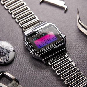 May include: A digital Casio watch with a silver metal band and a black face displaying the time. The watch face shows the time as 11:24:19 PM. Additional watch components and a gray watch strap are also visible.