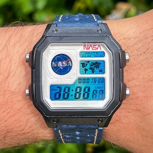 Könnte beinhalten: Eine schwarze Digitaluhr mit blauem Lederarmband wird gezeigt. Das Zifferblatt zeigt das NASA-Logo und die Uhrzeit an. Die Uhr hat ein quadratisches Zifferblatt mit weißem Hintergrund und blauen Digitalziffern. Das Armband ist blau mit weißen Nähten.