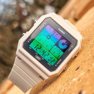 May include: A white Casio digital watch with a multi-coloured display showing the time as 12:42. The watch has a square face with a black bezel and a white strap.