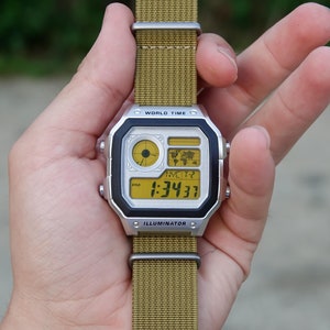 Könnte beinhalten: Eine silberne Digitaluhr mit schwarzem Zifferblatt und gelber digitaler Anzeige. Die Uhr hat eine schwarze und silberne Lünette und ein braunes Nylonarmband. Das Zifferblatt der Uhr zeigt die Zeit, das Datum und die Weltzeit an. Die Uhr verfügt außerdem über eine Leuchtfunktion.