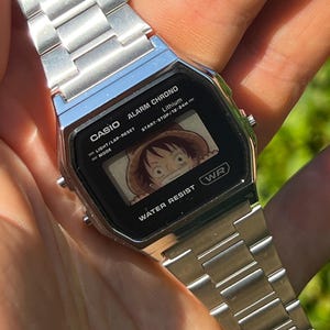 May include: A silver Casio digital watch with a black face showing a cartoon character. The watch has a stainless steel strap. The text "CASIO" and "WATER RESIST" are visible on the watch face.