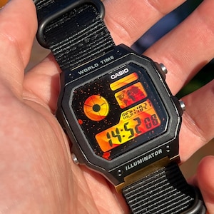 Op de afbeelding: Een zwart Casio-horloge met een zwarte nylon band en een gouden bezel. De wijzerplaat van het horloge is zwart met een rood en oranje digitaal display. Het horloge heeft een wereldtijdfunctie en een verlichting.