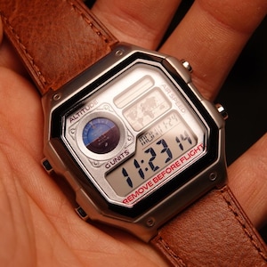 May include: A digital watch with a silver case and a brown leather strap. The watch face displays the time, altitude, and other data. The text "REMOVE BEFORE FLIGHT" is visible on the watch face.