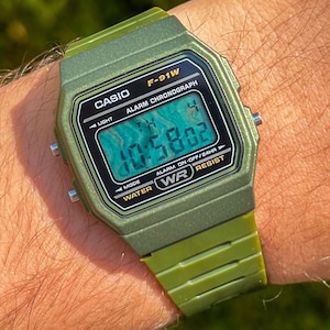 Puede incluir: Un reloj digital Casio F-91W verde se muestra en una muñeca. El reloj tiene una esfera rectangular con una pantalla digital que muestra la hora. La correa y la caja del reloj son de color verde a juego. El reloj es resistente al agua.