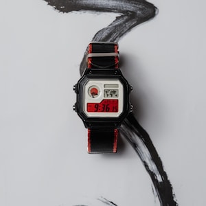 Può includere: Orologio digitale nero con cinturino in nylon rosso e nero. Il quadrante mostra l'ora 9:36 e presenta una grafica del sole rossa. L'orologio è posto su uno sfondo bianco con un design a pennellate nere.